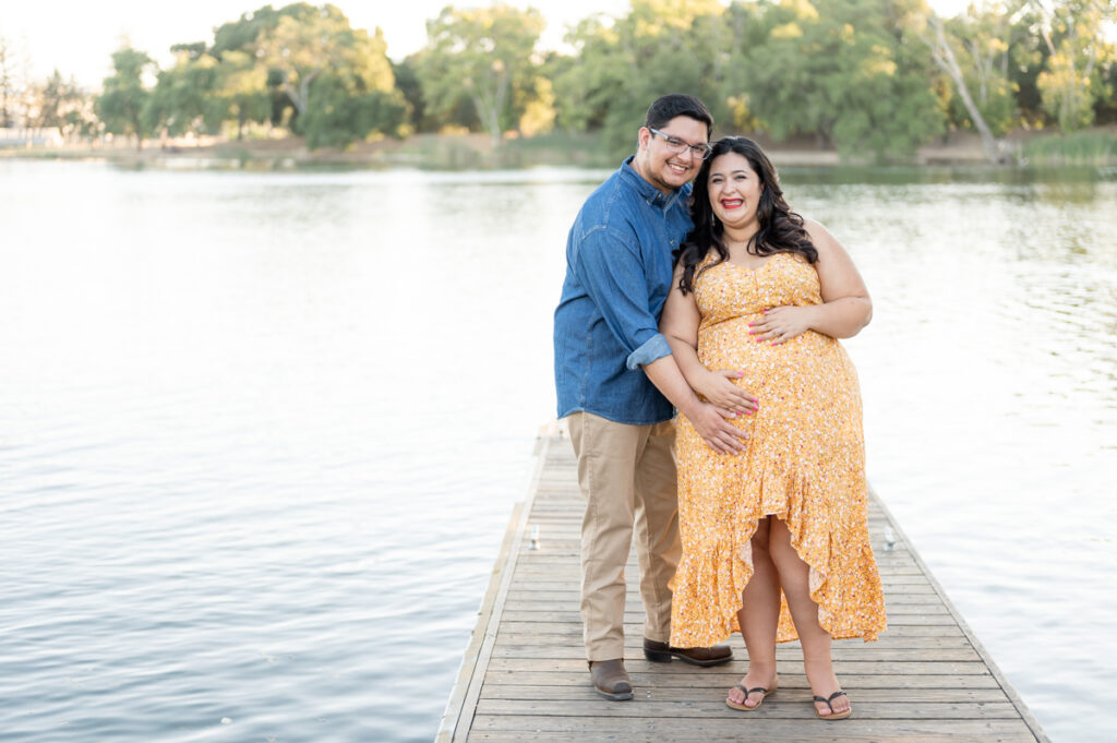 Dad and pregnant mom posing on a dock near the water during a maternity session at Lodi Lake Park, Lodi CA with Gia Chong Photography, Lodi Photographer