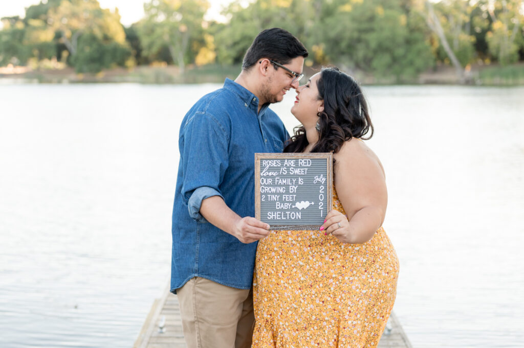 Parents holding a letter board announcement on a dock near the water during a maternity session at Lodi Lake Park, Lodi CA with Gia Chong Photography, Lodi Photographer