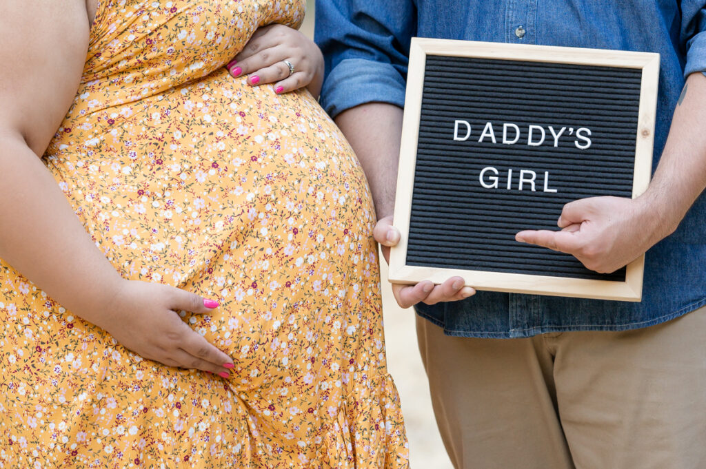 Dad and pregnant mom posing with a letter board in a field during a maternity session at Lodi Lake Park, Lodi CA with Gia Chong Photography, Lodi Photographer