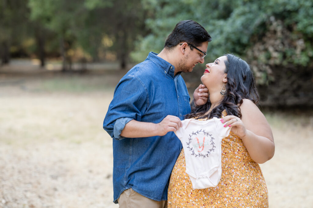 Dad and pregnant mom posing with ta baby onesie in a field during a maternity session at Lodi Lake Park, Lodi CA with Gia Chong Photography, Lodi Photographer