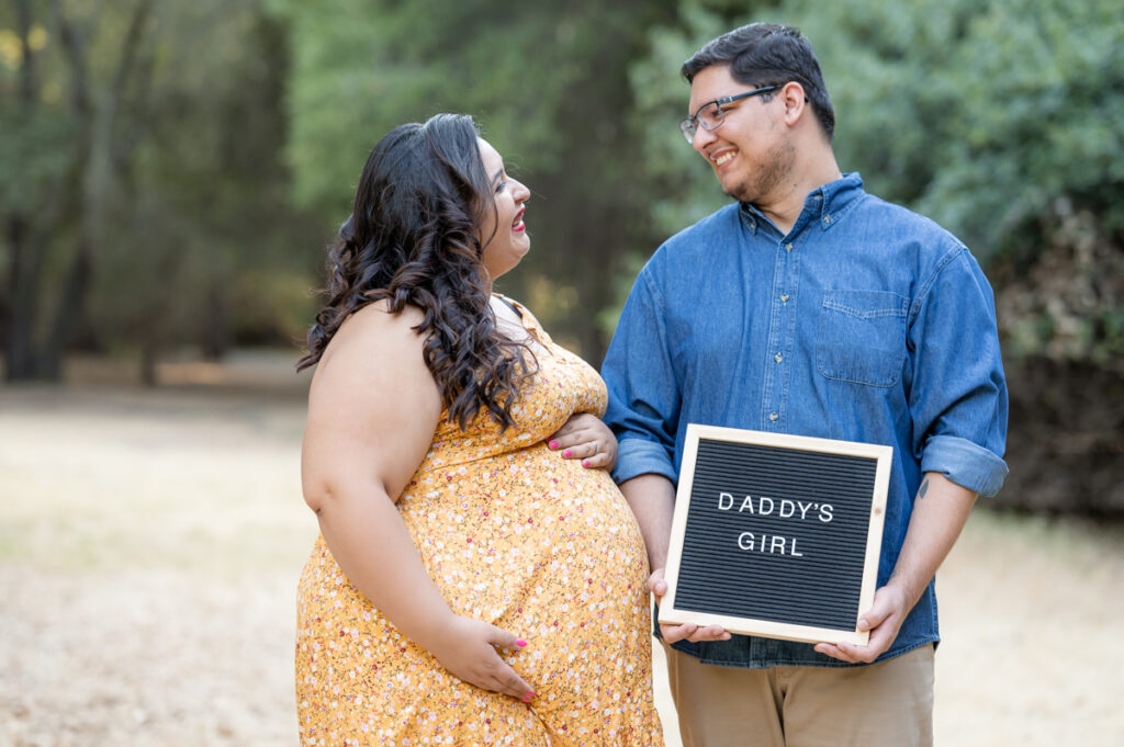 Dad and pregnant mom posing with a letter board in a field during a maternity session at Lodi Lake Park, Lodi CA with Gia Chong Photography, Lodi Photographer