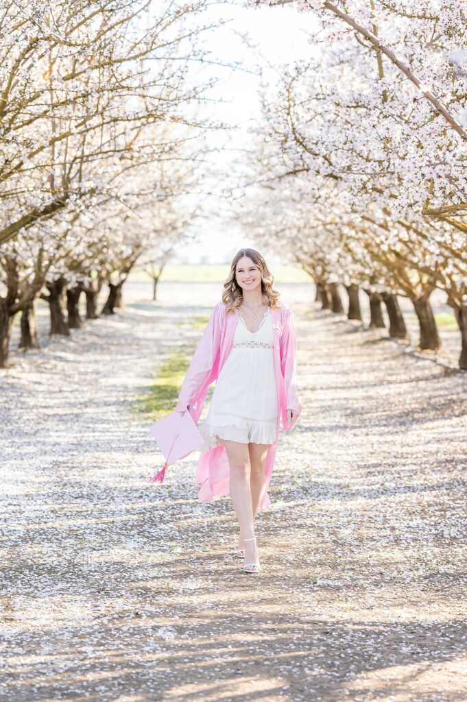 senior grad walking in a white dress and grad gown in almond blossoms session