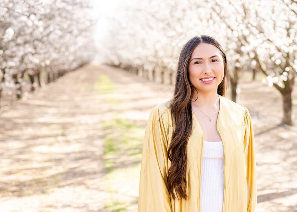 Senior girl in grad gown smiling at camera during almond blossom grad session